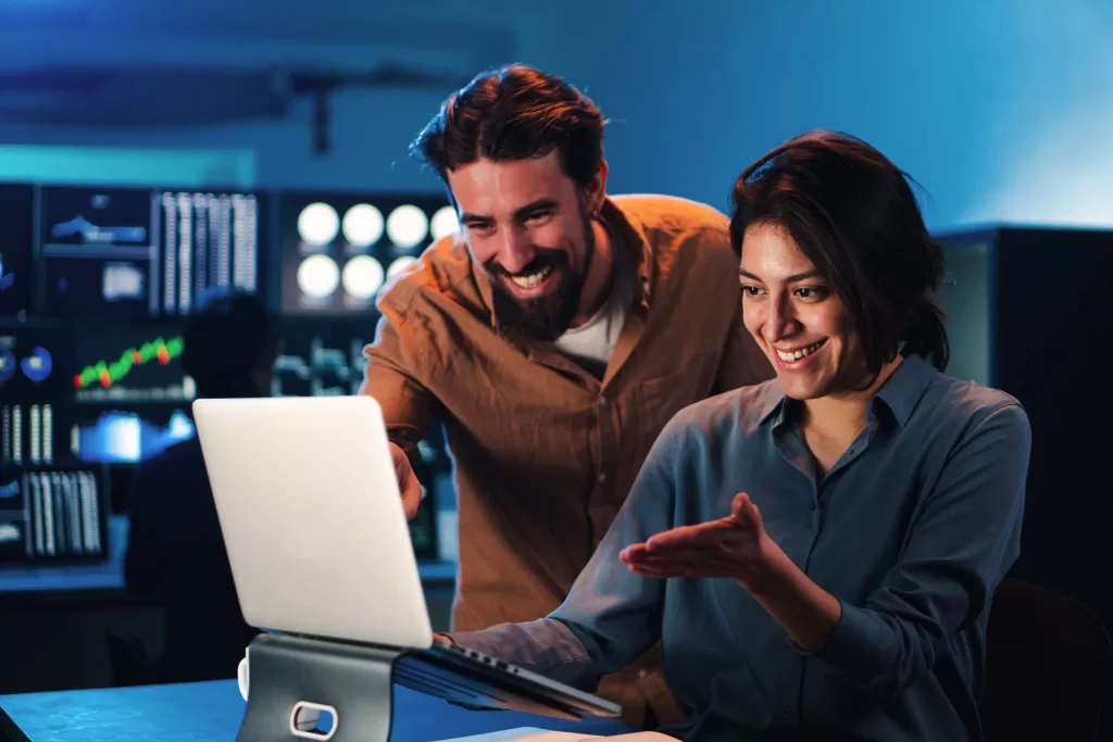 Happy business couple working on a laptop computer in the office. Young trading team people smiling watching data on pc screen. Two partners coworking on finance. High quality photo