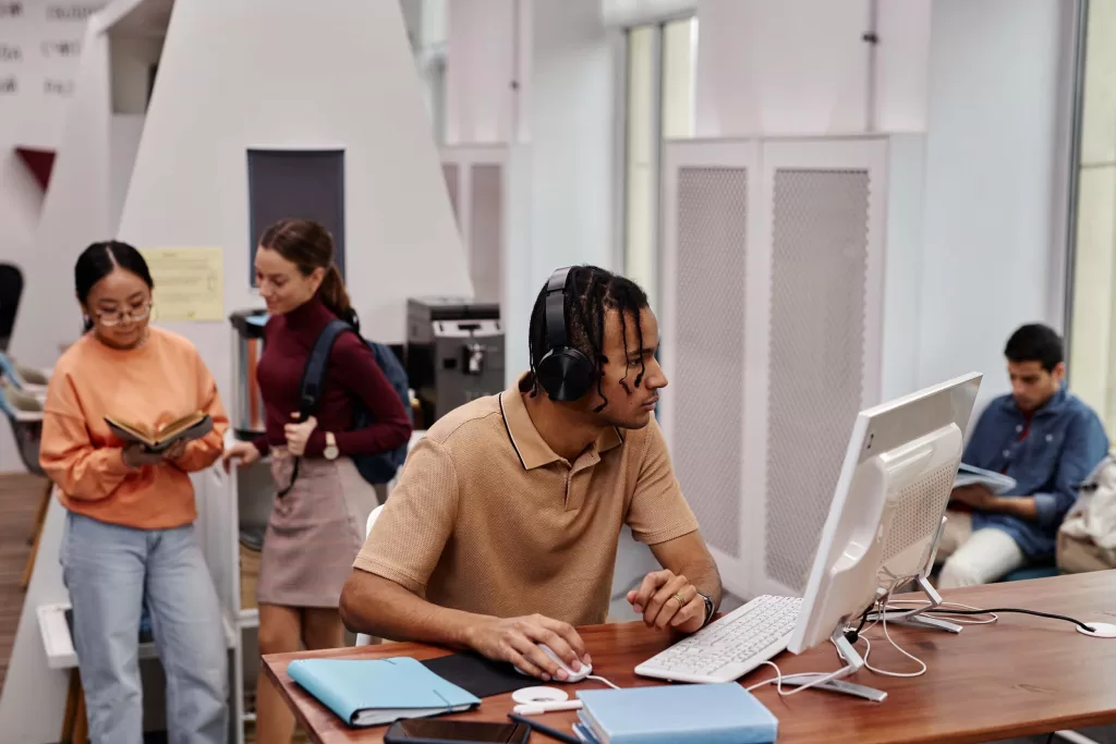 Side view portrait of young black man using computer in college library and wearing headphones with people in background