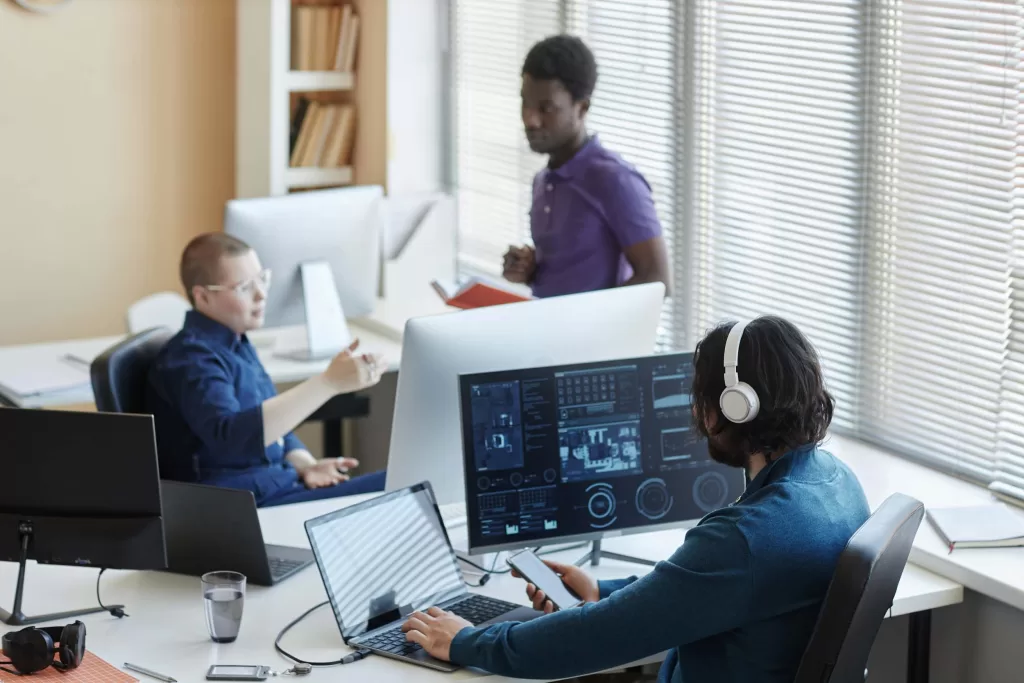 Young worker of cyber security center sitting in front of computers with graphic data and using smartphone while his colleagues having discussion